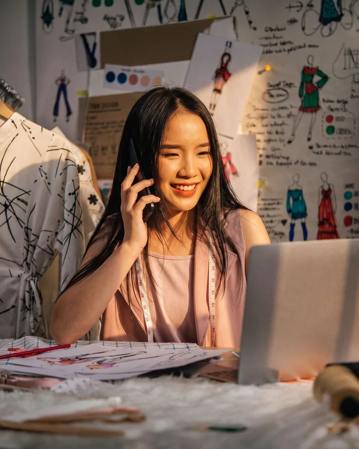 A fashion designer on the phone at her laptop, surrounded by sketches and fabric
